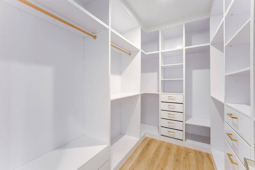 This large custom built closet  features built-in drawers with gold  accented hardware, storage space,  and clothes hanging rods.
