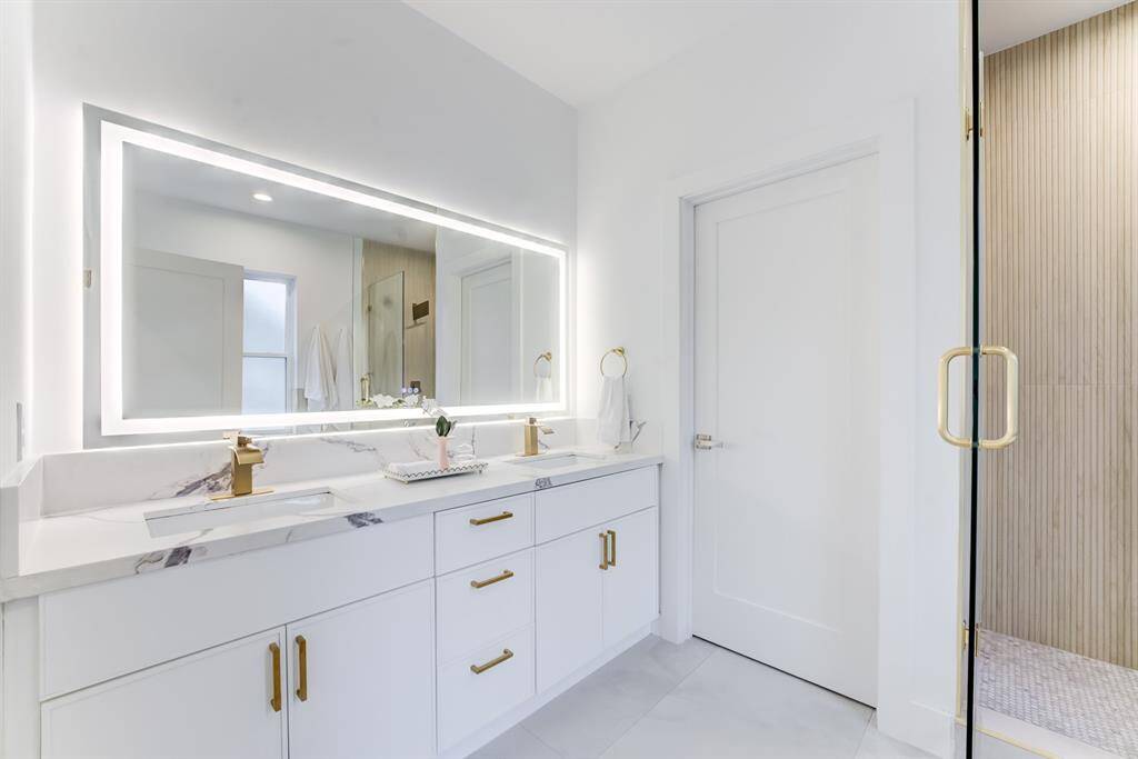 The ensuite bathroom has a large  lighted mirror, dual sinks with gold  hardware, quartz countertops, and  sleek white cabinetry for storage.