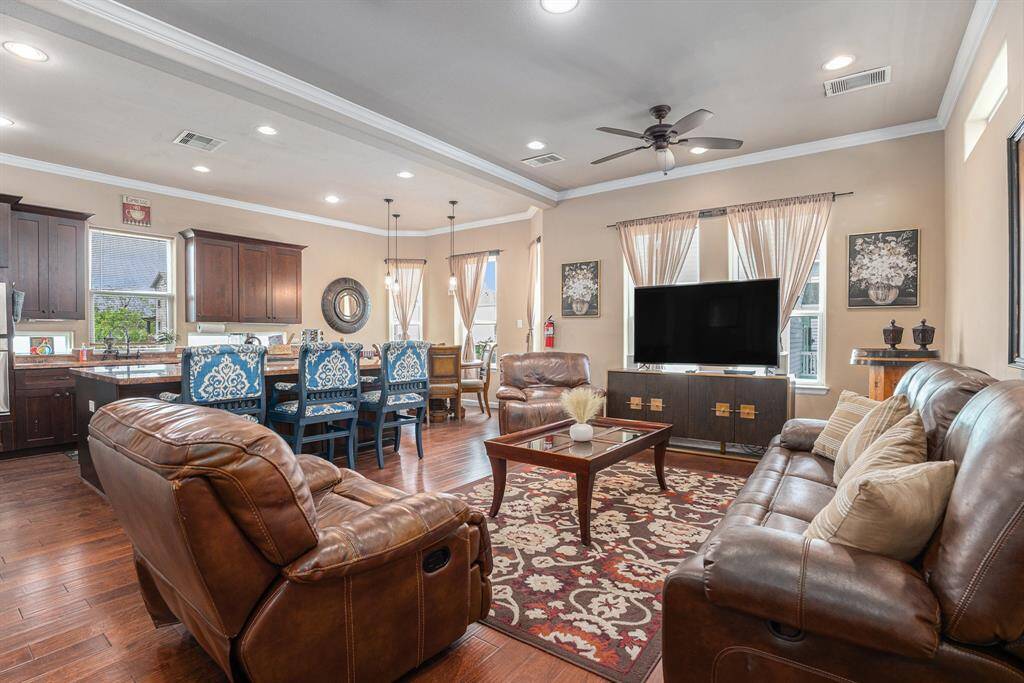 The second-floor family room offers a refined open layout with hardwood floors, a leather sofa and recliner, a patterned rug, a wooden coffee table, a large TV on a modern console, a dining area with blue upholstered chairs, and a kitchen island, all enhanced by recessed lighting and natural light from curtained windows.