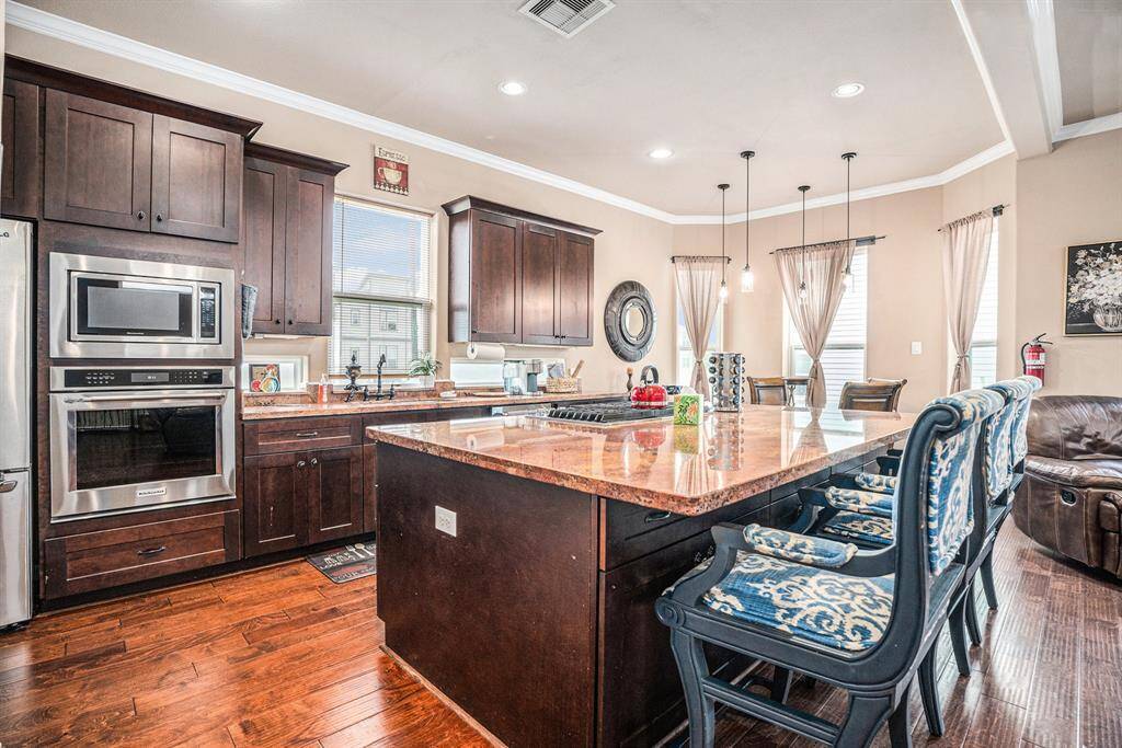 A modern kitchen showcases rich wooden cabinetry, a large granite-topped island with blue patterned bar stools, stainless steel appliances, and pendant lighting, seamlessly connecting to an open dining and living area with hardwood floors and tasteful decor.