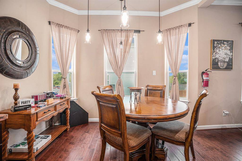 The breakfast area features a round wooden table with four cushioned chairs, illuminated by pendant lights, and is complemented by a wooden sideboard, a large round mirror, a floral painting, and three windows with sheer curtains.