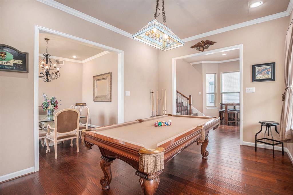 Adjacent to the dining room and near the balcony, this pool room features a sleek wooden billiards table under a contemporary stained-glass light, complemented by hardwood floors, neutral decor, and ample natural light from large windows with blinds.