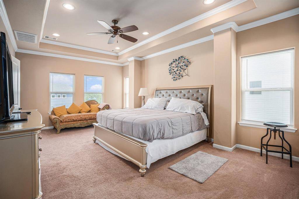 The primary bedroom provides a comfortable retreat with a tufted headboard bed, a plush gray comforter, a mustard-yellow chaise lounge with pillows, a white dresser, a ceiling fan, a tray ceiling with recessed lighting, and two structural columns, all complemented by two windows with blinds on soft carpeted flooring.