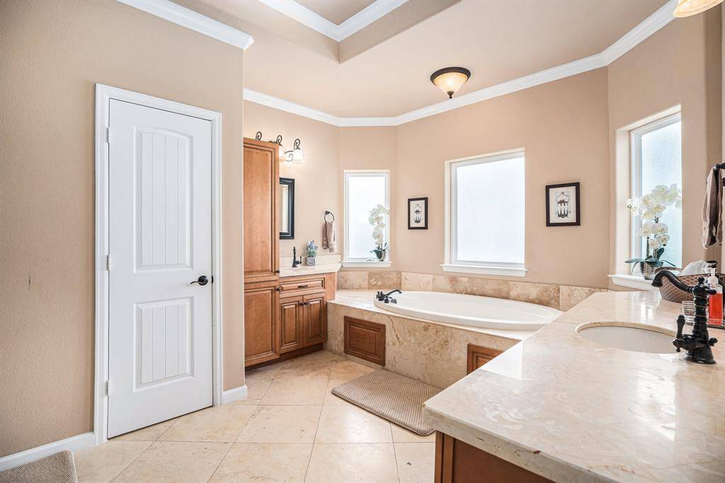 The primary bathroom includes a large soaking tub under three frosted glass windows, two separated sinks on a marble countertop, wooden cabinetry, a tiled floor, and tray ceilings, all lit by a ceiling light and wall sconces.