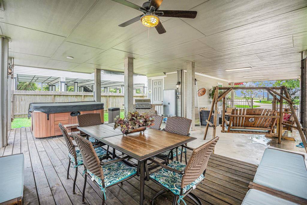 This expansive covered patio, professionally designed with a wooden dining table, wicker chairs, a hot tub, a built-in mobile grill, and a wooden swing, provides an elegant outdoor living space within a well-maintained, fenced backyard.