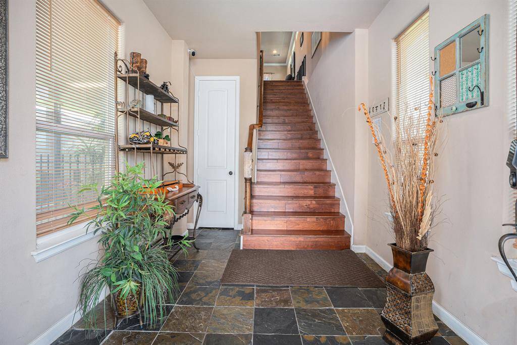This elegantly designed first-floor foyer features a wooden staircase, slate tile flooring, a decorative console table with shelves, a potted plant, and a tall vase with dried branches, accented by natural light from two windows with blinds.