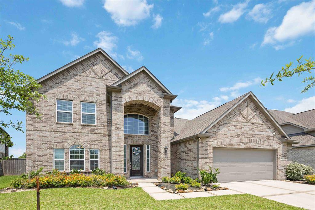 This photo showcases a two-story brick house with a gabled roof and a two-car garage. The home features large windows, a welcoming entryway with an arched window above, and a well-maintained front yard with landscaping. The sky is clear and blue, enhancing the property's curb appeal.