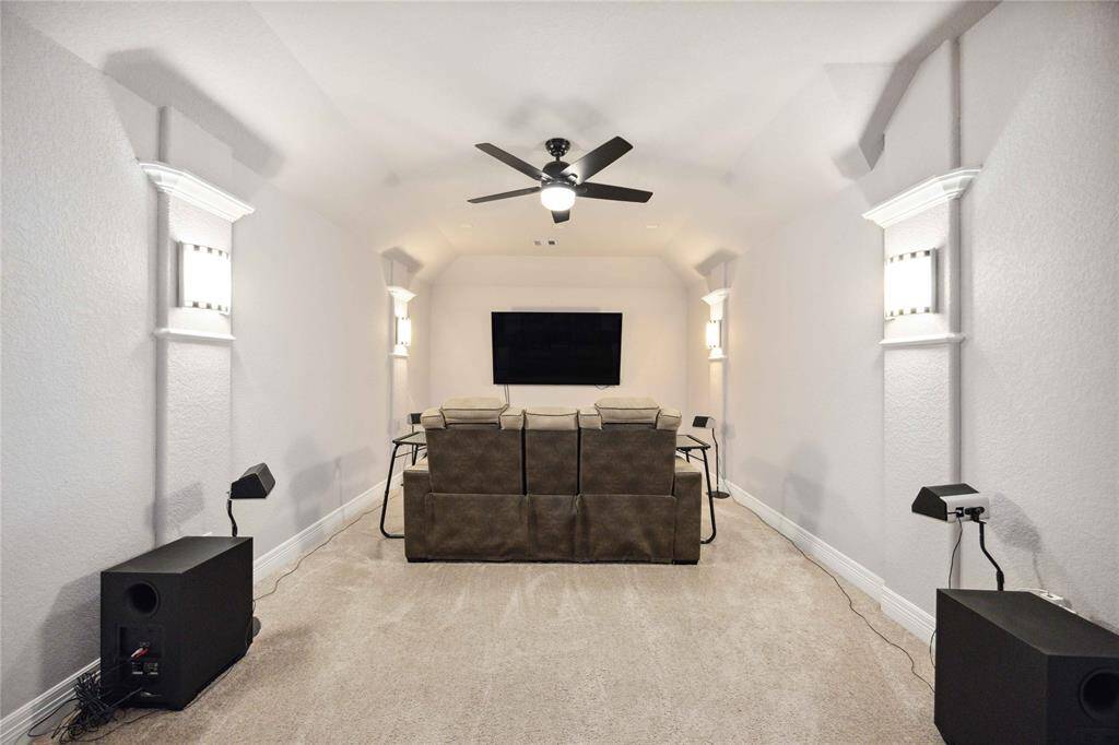 This photo shows a cozy home theater room with comfortable seating, a large wall-mounted TV, and ambient wall lighting. The space is carpeted, featuring a ceiling fan and surround sound speakers, making it perfect for entertainment.