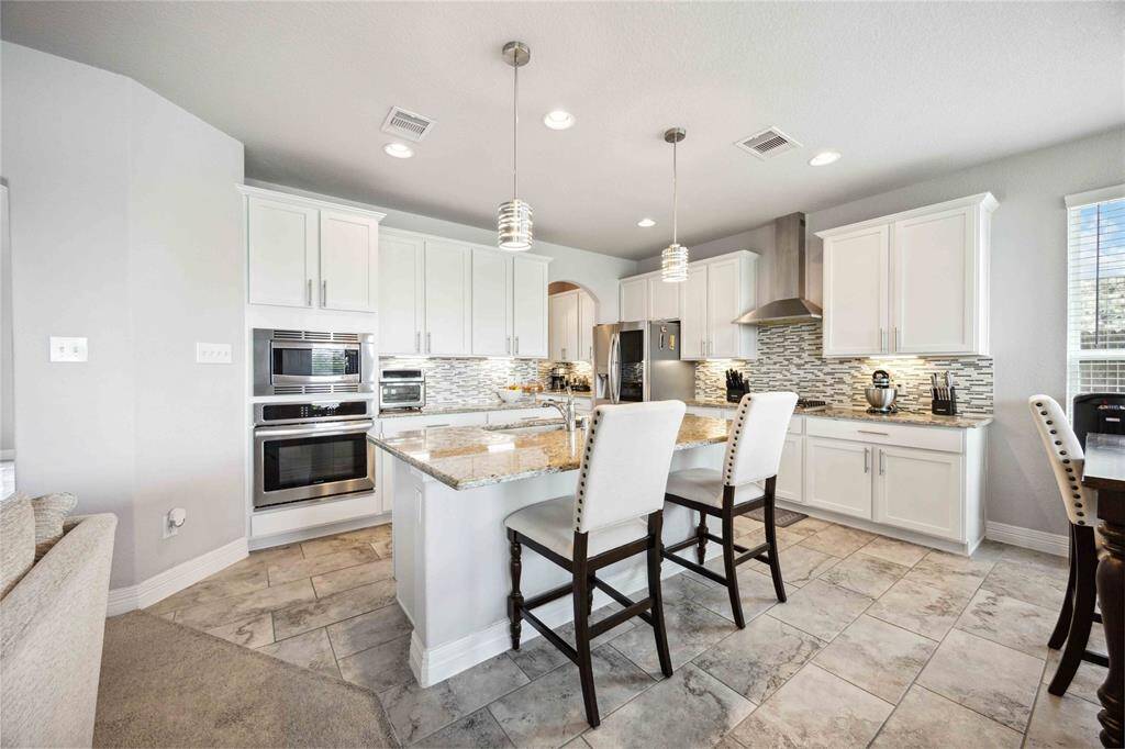 This modern kitchen features sleek cabinetry, a stylish tile backsplash, and stainless-steel appliances. An island with granite countertops offers seating and pendant lighting, creating a bright and inviting space.