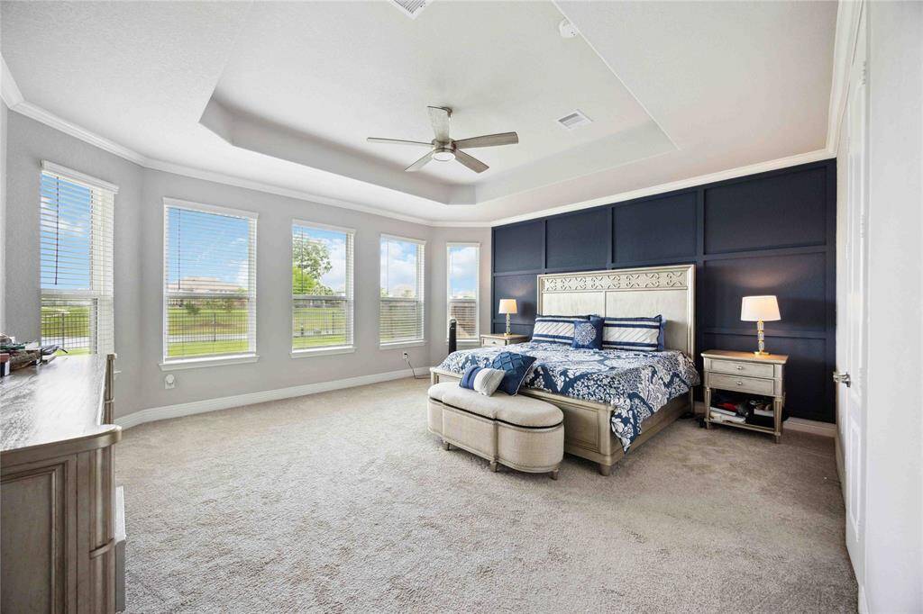 This spacious primary bedroom features large windows allowing ample natural light, a stylish tray ceiling with a fan, and a modern accent wall, creating an inviting and serene atmosphere.