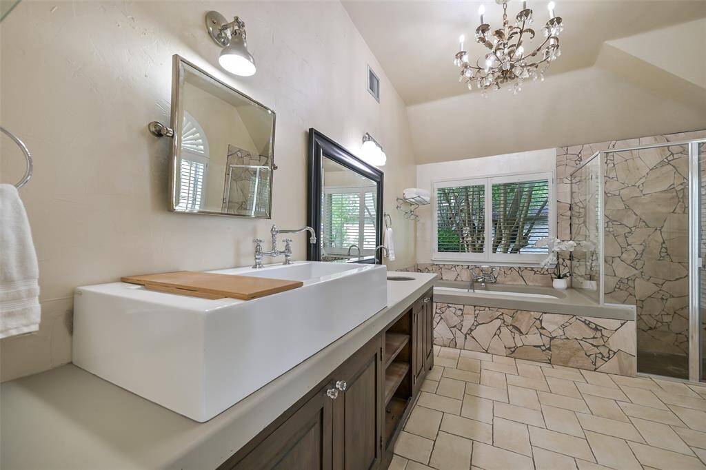 The spa-like primary bath showcases a dramatic chandelier, dual sink vanity with a farmhouse-style basin, a bubbler tub for relaxing, and a large walk-in shower surrounded by striking natural stone tilework.