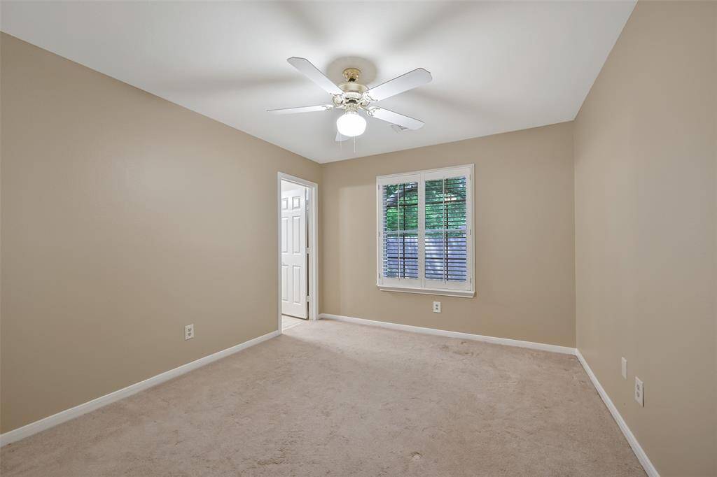 This comfortable secondary bedroom features neutral tones, a side-facing window shaded by plantation shutters, a lighted ceiling fan, and a private en suite bath—ideal for guests or multi-generational living.