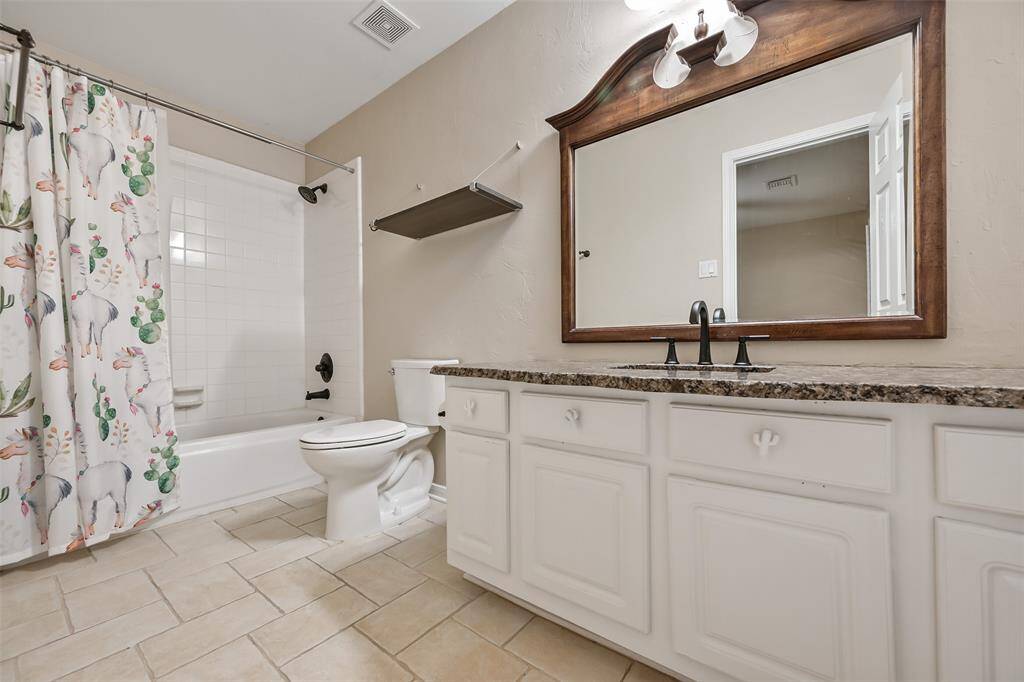 The private en suite bath features a raw edge granite-topped vanity with an undermount stainless steel sink, oil-rubbed bronze fixtures, white cabientry, a framed mirror, tile floors, and a classic tub/shower combo with crisp white tile surround.