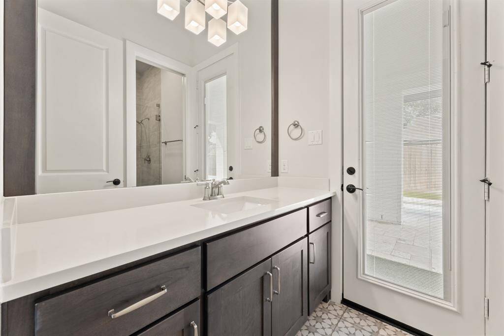 Modern bathroom with sleek dark cabinetry, a large mirror, and a stylish light fixture. Features a glass door leading outside, offering natural light and easy access.