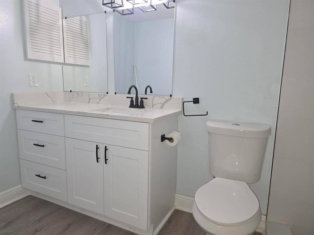 This is a modern bathroom featuring a sleek white vanity with black fixtures, a large mirror, and a light fixture above. The room has light-colored walls and wood-style flooring, creating a clean and contemporary look.