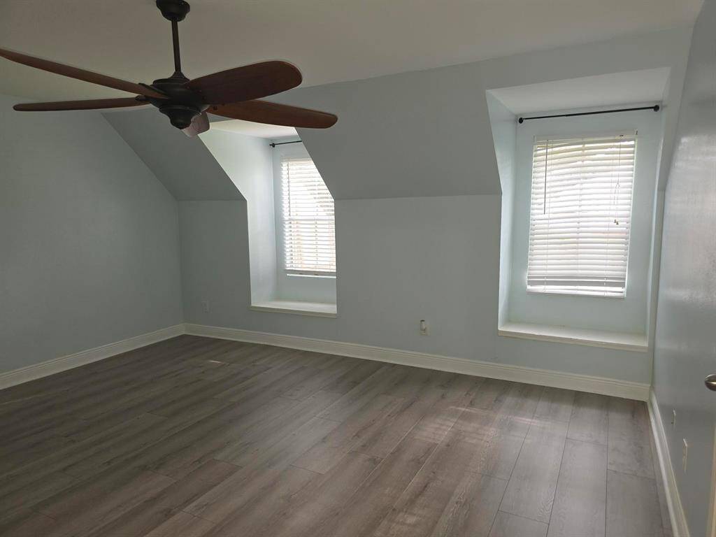 This room features light gray walls and hardwood floors, with two large windows that provide ample natural light. It has a ceiling fan and a clean, modern aesthetic, ideal for a bedroom or office space.