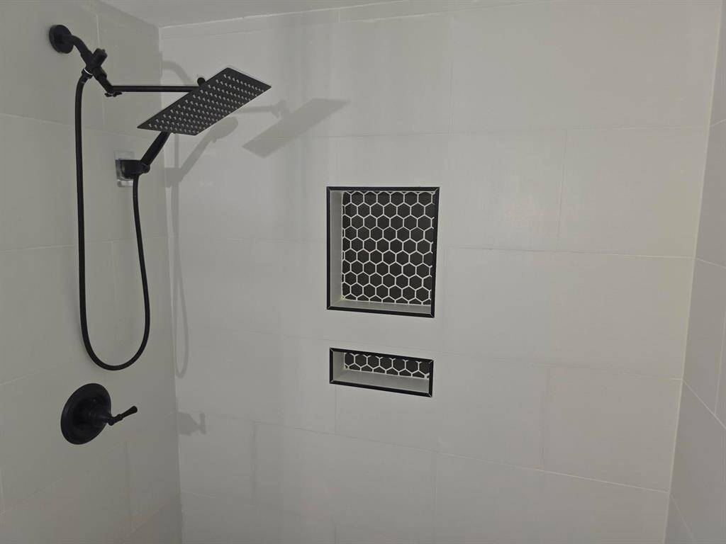 This photo shows a modern shower with a sleek matte black rain shower head and fixtures. It features a white tiled wall with two built-in niches, accented by black hexagonal tile designs.