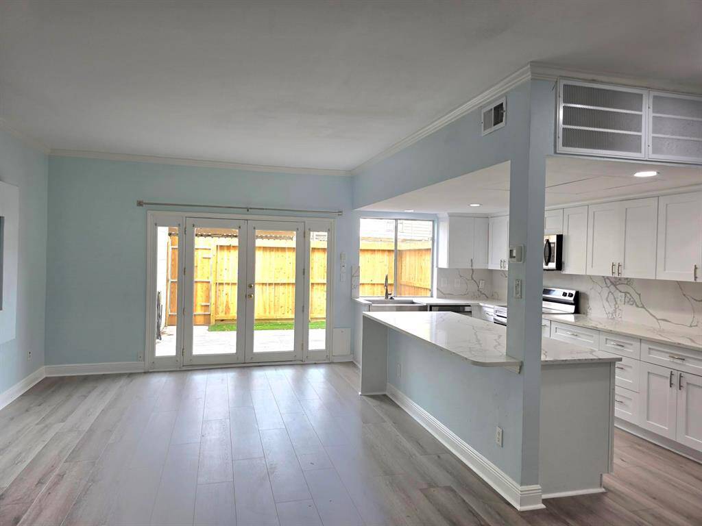 This photo showcases a modern open-plan kitchen and living area. It features sleek stainless steel appliances, a large island with marble countertops, and light wood flooring. The space is brightened by natural light from large windows and a sliding glass door leading to a fenced yard.