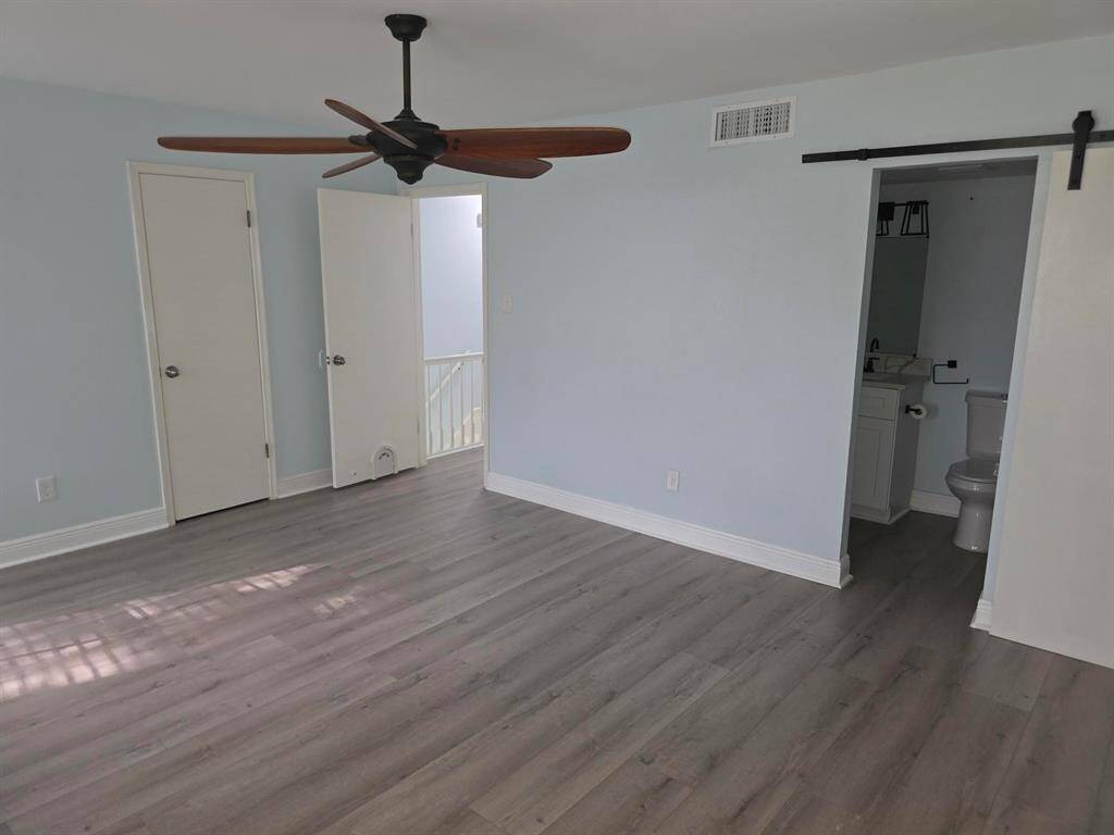 This room features light blue walls, laminate wood flooring, and a ceiling fan. It has an en-suite bathroom with a sliding barn door and is well-lit with natural light.