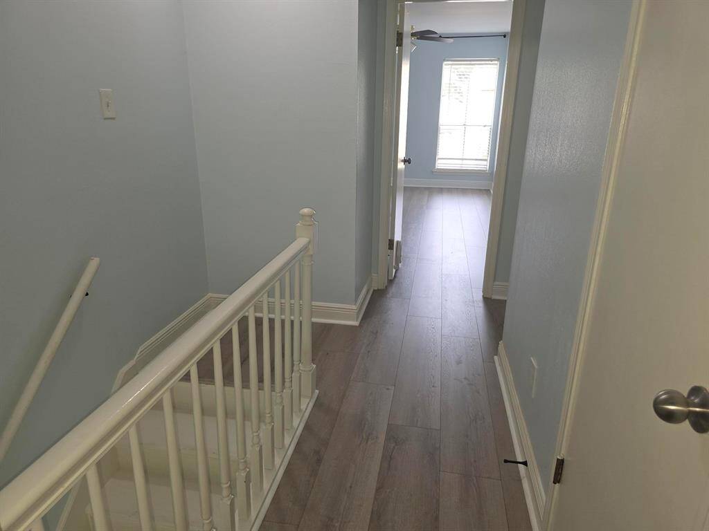 This photo shows a bright, airy hallway with light-colored walls and flooring. It features a staircase with a white railing, leading to a room with a window and ceiling fan, allowing natural light to flow in.