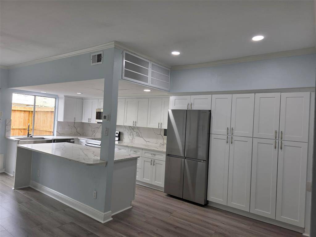 This kitchen features modern white cabinetry, a sleek stainless steel refrigerator, and a spacious marble-patterned countertop. It's well-lit with recessed lighting and offers a bright, open layout with a large window for natural light.