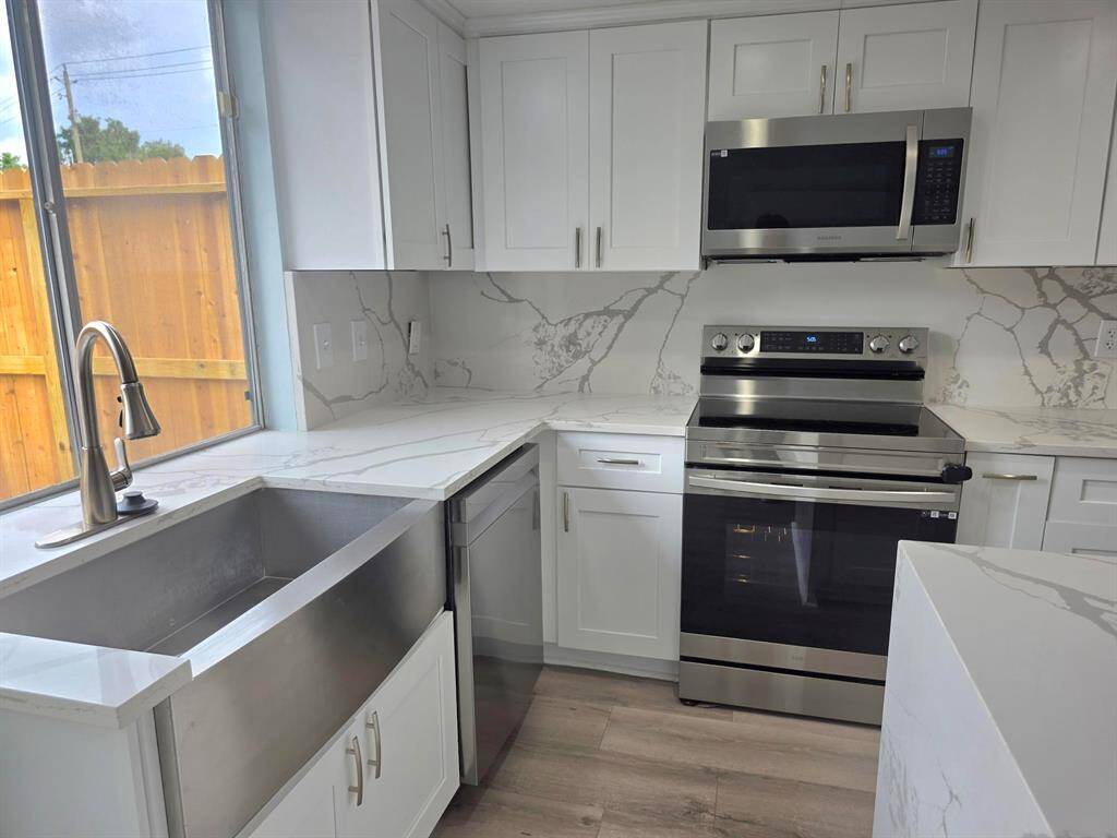 This photo showcases a modern open-plan kitchen and living area. It features sleek stainless steel appliances, a large island with marble countertops, and light wood flooring. The space is brightened by natural light from large windows and a sliding glass door leading to a fenced yard.