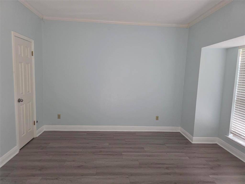 This room features light blue walls and sleek gray wood flooring. It has a door on the left and a large window with blinds on the right, providing plenty of natural light. The space is simple and versatile, ideal for personalizing.
