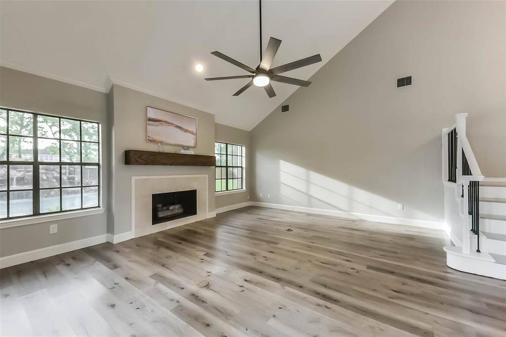 This spacious living room features high vaulted ceilings, a modern ceiling fan, and large windows looking out to the amazing backyard. It features a cozy fireplace with a wooden mantel and light wood flooring, creating a warm and inviting atmosphere. Virtually staged
