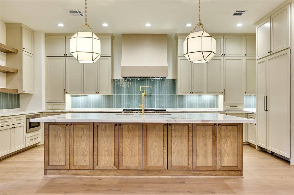 This kitchen features a spacious island with a wood finish, elegant pendant lighting, and ample cabinetry for storage. The design includes a modern backsplash, high-end appliances, and a bright, open layout. Perfect for cooking and entertaining.