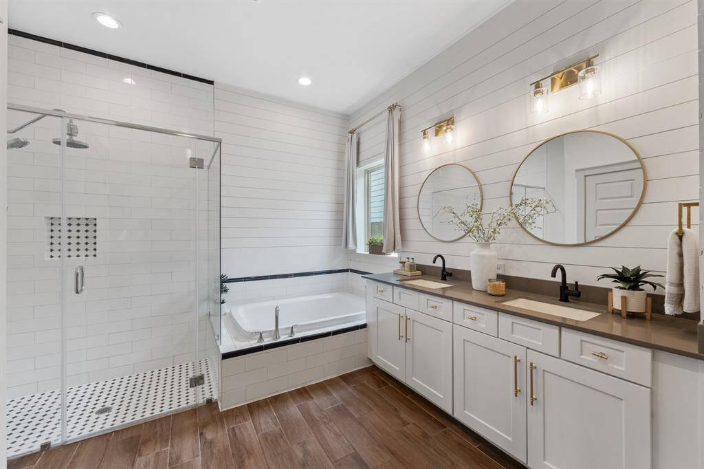 The primary bathroom boasts black faucets and double sinks atop brown glass countertops, complemented by white cabinets and gold-framed circular mirrors for a modern, luxurious aesthetic.