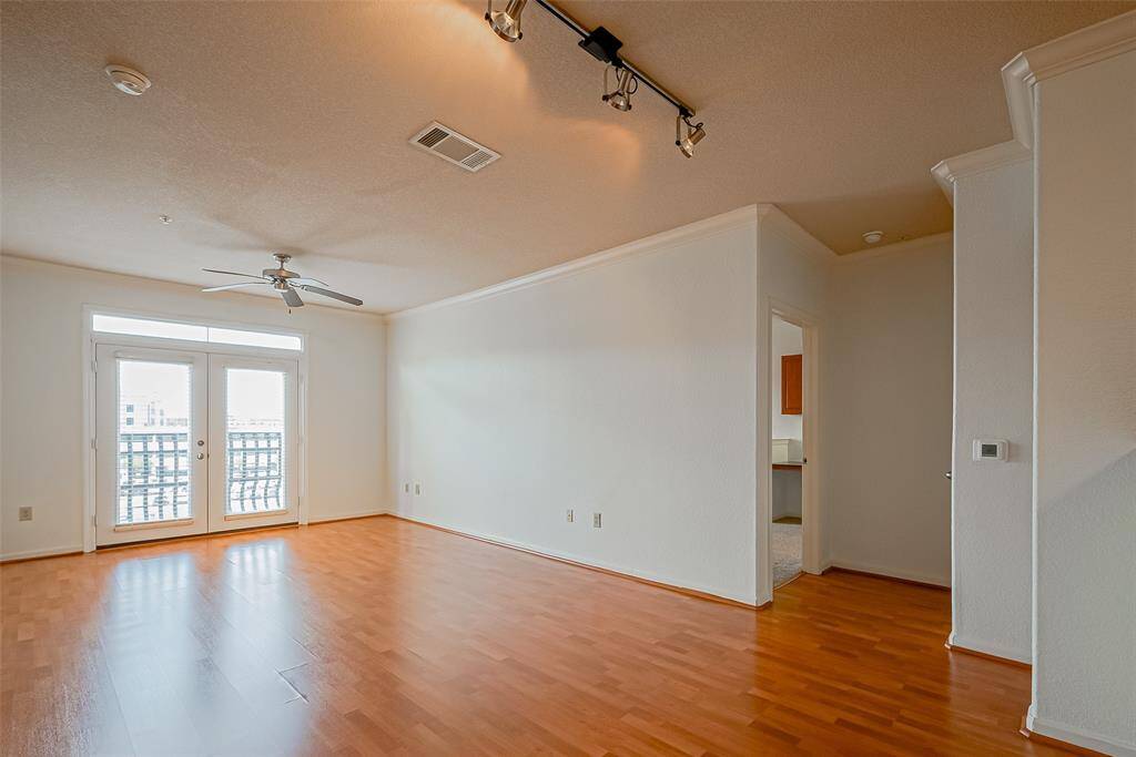 This spacious living area features laminate floors, a ceiling fan, and track lighting. It has large French doors leading to a balcony, providing plenty of natural light. The open layout connects to the kitchen area, enhancing the flow of the space.