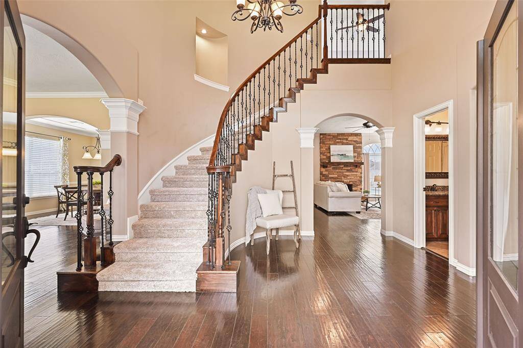 The newly installed double doors open into a grand foyer with a soaring 18-foot ceiling with a scrolled iron chandelier, hand-scraped engineered wood flooring, fresh neutral paint, and a sweeping staircase with carpeted treads, wood handrails, and wrought iron basketweave balusters.