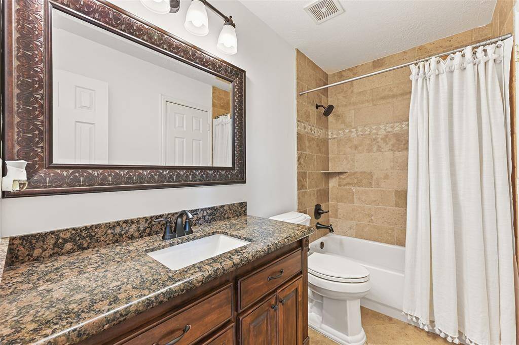 This full bathroom is accessible from the foyer or the study and it features a granite top vanity with a single rectangular basin sink, stained wood cabinetry, a framed mirror, stylish lighting, tile floors, and a combination bathtub and shower with a tile surround.