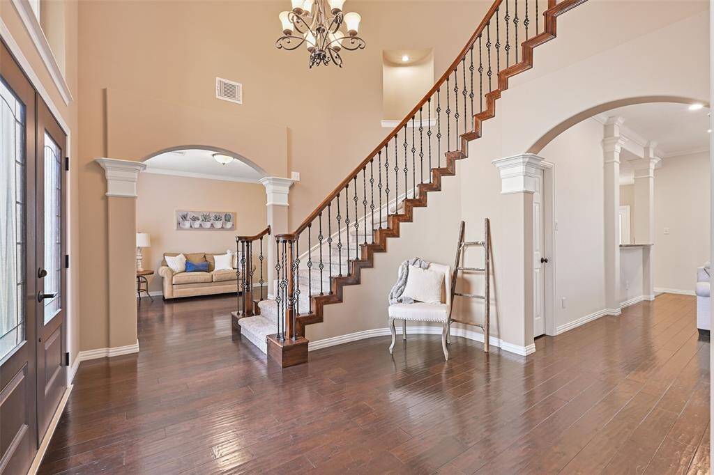 The newly installed double doors open into a grand foyer with a soaring 18-foot ceiling with a scrolled iron chandelier, hand-scraped engineered wood flooring, fresh neutral paint, and a sweeping staircase with carpeted treads, wood handrails, and wrought iron basketweave balusters.