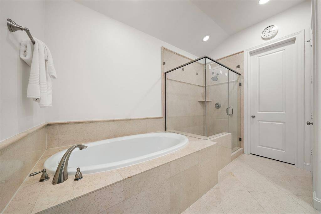 The ensuite bath features a wide soaking tub and glass-enclosed shower wrapped in neutral stone tile—an inviting combination designed for both everyday ease and quiet moments of rest.