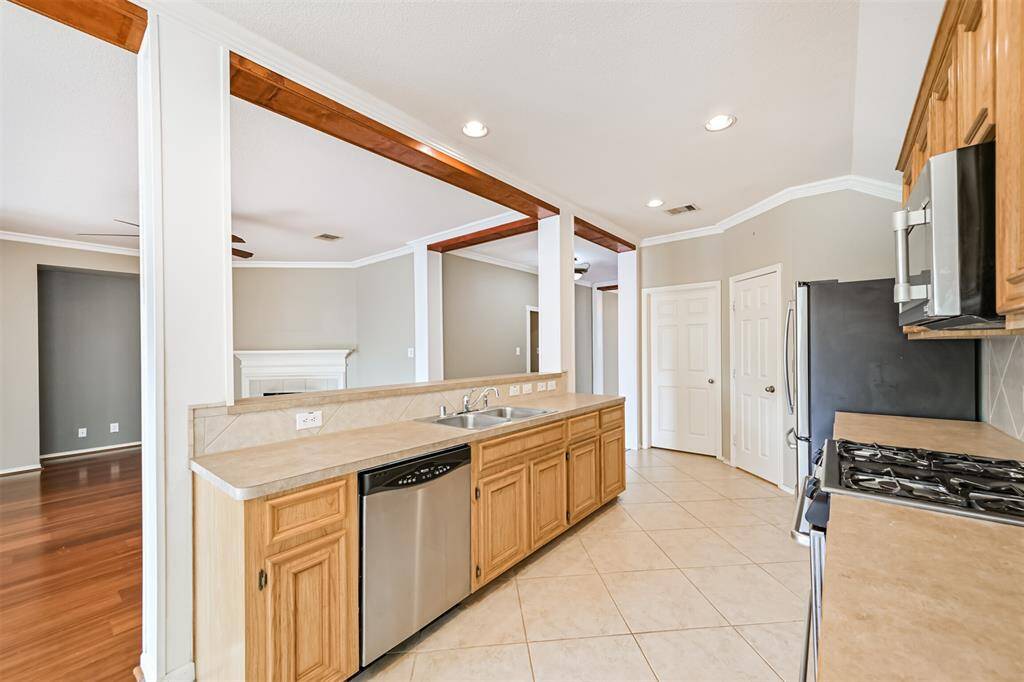 Spacious, open-concept kitchen and living area. The kitchen features wooden cabinets, a gas stove, and a double sink, with a dining area visible in the background. The space is bright with natural light, tile flooring, and a ceiling fan in the living area.