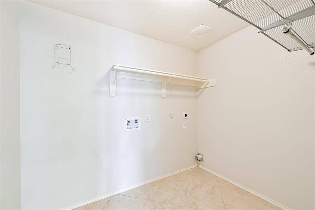 Bright and functional laundry room with tiled flooring, featuring a utility hook-up, wall-mounted shelf, and overhead metal shelving for ample storage.