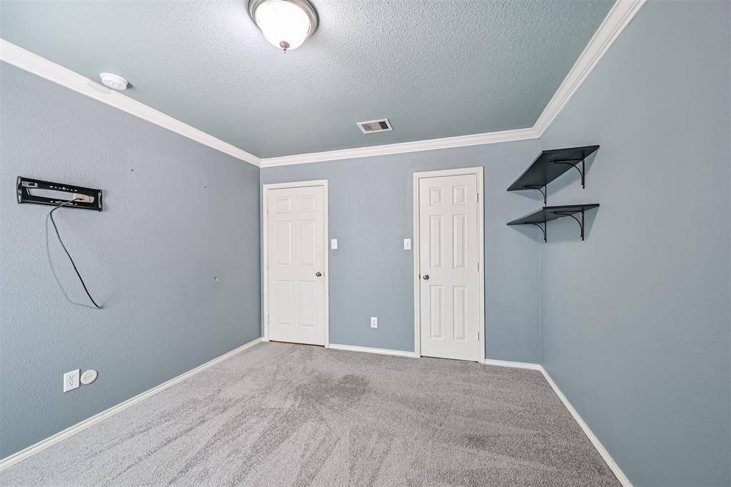 Guest bedroom with soft blue walls, crown molding, and light carpet. It features a ceiling light fixture, built-in shelving, and two doors, one leading to a hallway.