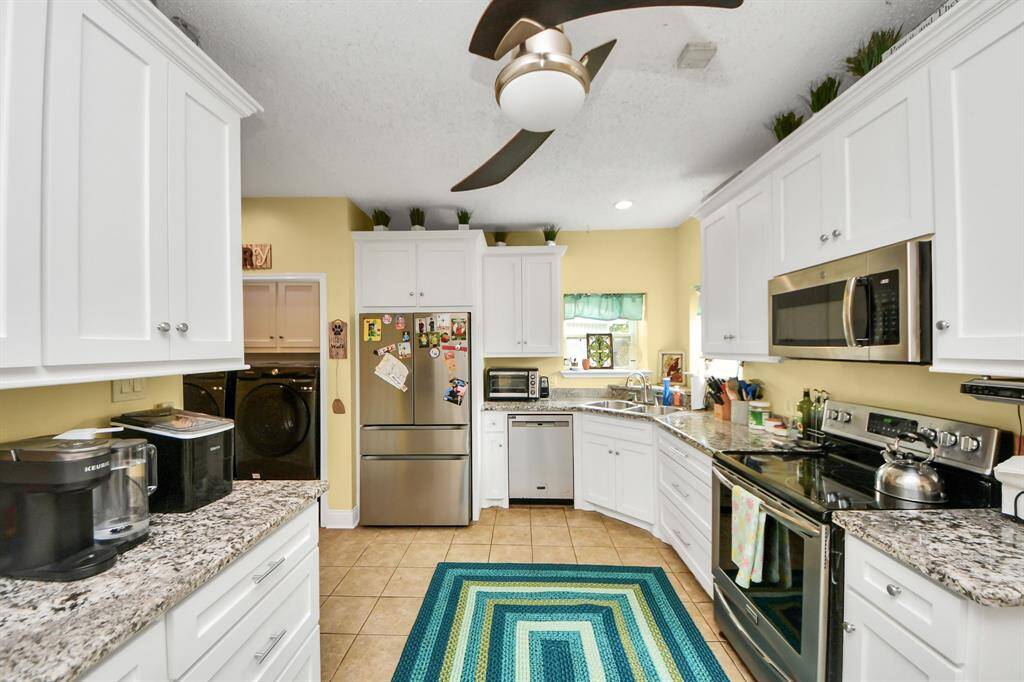 This kitchen features modern stainless steel appliances, white cabinetry, and granite countertops. The space is well-lit with natural light and has a convenient adjacent laundry area.