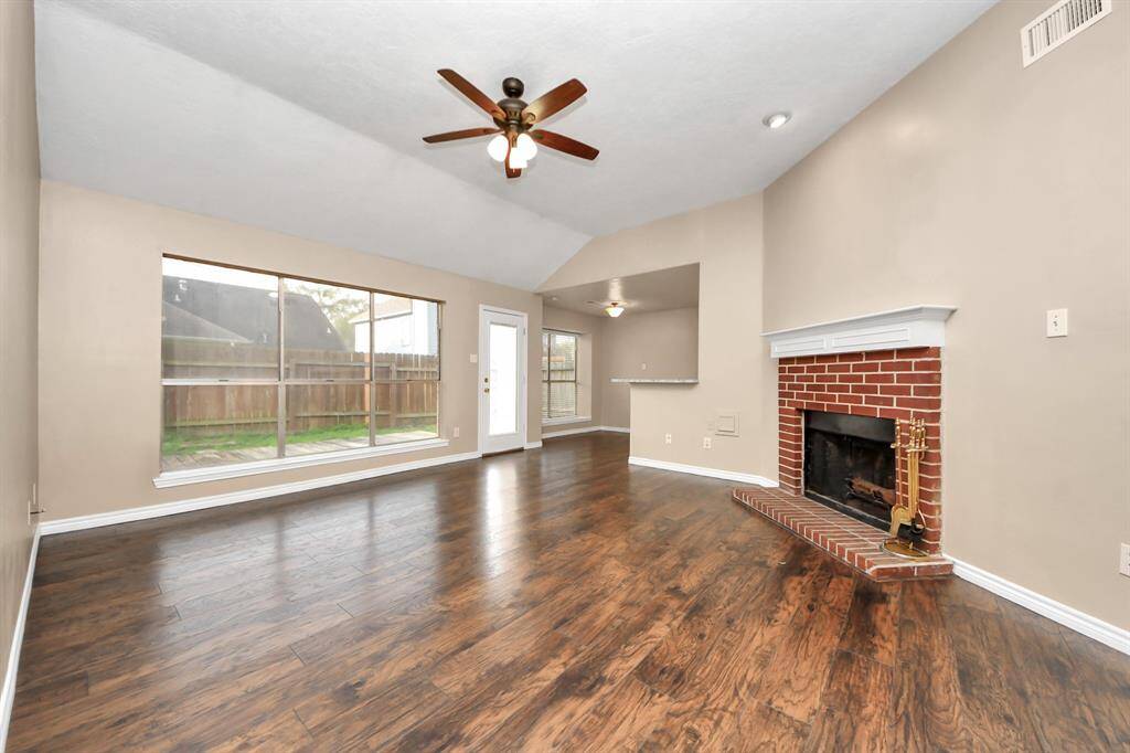 This spacious living room features hardwood floors, a cozy brick fireplace, large windows for natural light, and a ceiling fan. It opens to an outdoor view and connects seamlessly to the dining area.