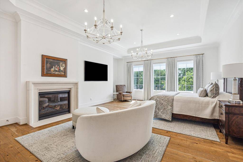 This serene primary suite  is a perfect retreat with its oversized windows, elegant lighting and drapes, handsome fireplace and space for various room configurations