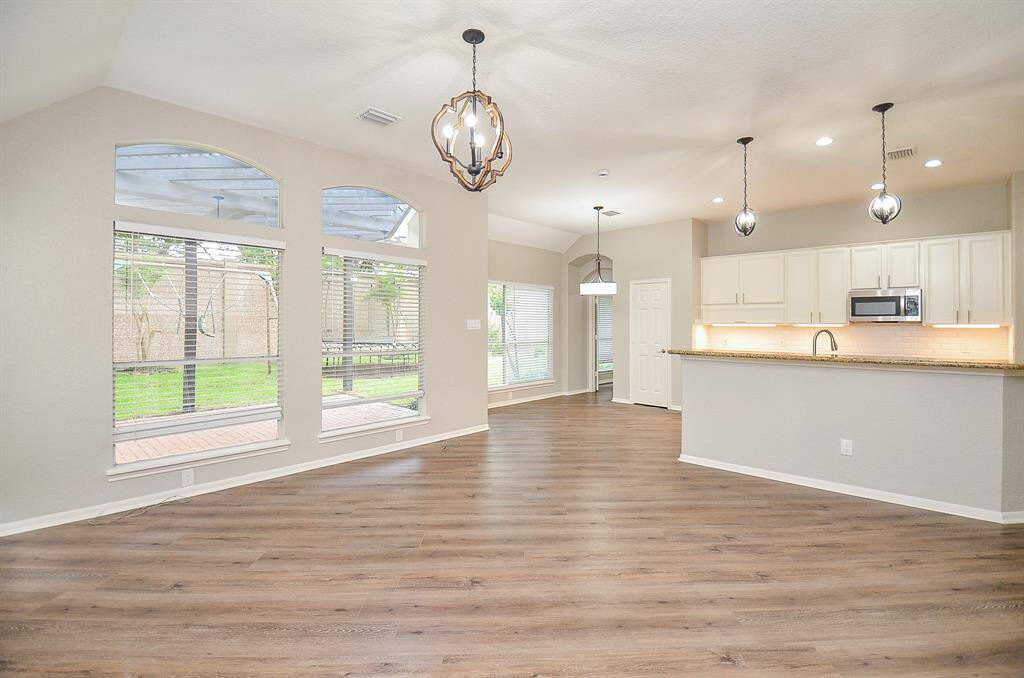 This view of the combined living/kitchen has a magical vibe! Wide open, airy space, up and down, defines the timeless beauty and versatility of the open floor plan design concept. Walls of huge arched windows overlook patio/backyard, with gorgeous pendant lighting embracing kitchen work surfaces, and a charming chandelier warming the breakfast/casual dining nook, up center.
