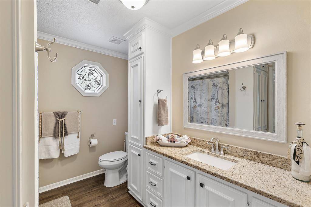 This bathroom features elegant beige walls, a granite countertop, and white cabinetry. It includes a large mirror with overhead lighting, a decorative octagonal window, and a shower. The space is well-lit and modern with wood-look flooring.