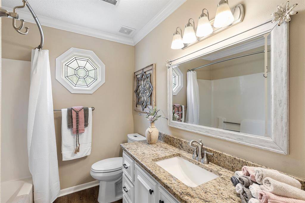 This secondary bathroom features a granite countertop with a stylish sink and faucet. It has a large framed mirror, modern light fixtures, and a bathtub with a shower curtain. The space is brightened by a unique octagonal window and decorated with tasteful accents, providing a cozy and inviting atmosphere.