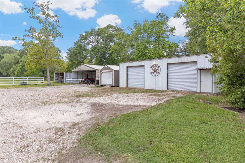 A spacious rural property with a large workshop featuring two roll-up garage doors. The area includes a gravel driveway, lush greenery, and a fenced-in pasture, perfect for country living.