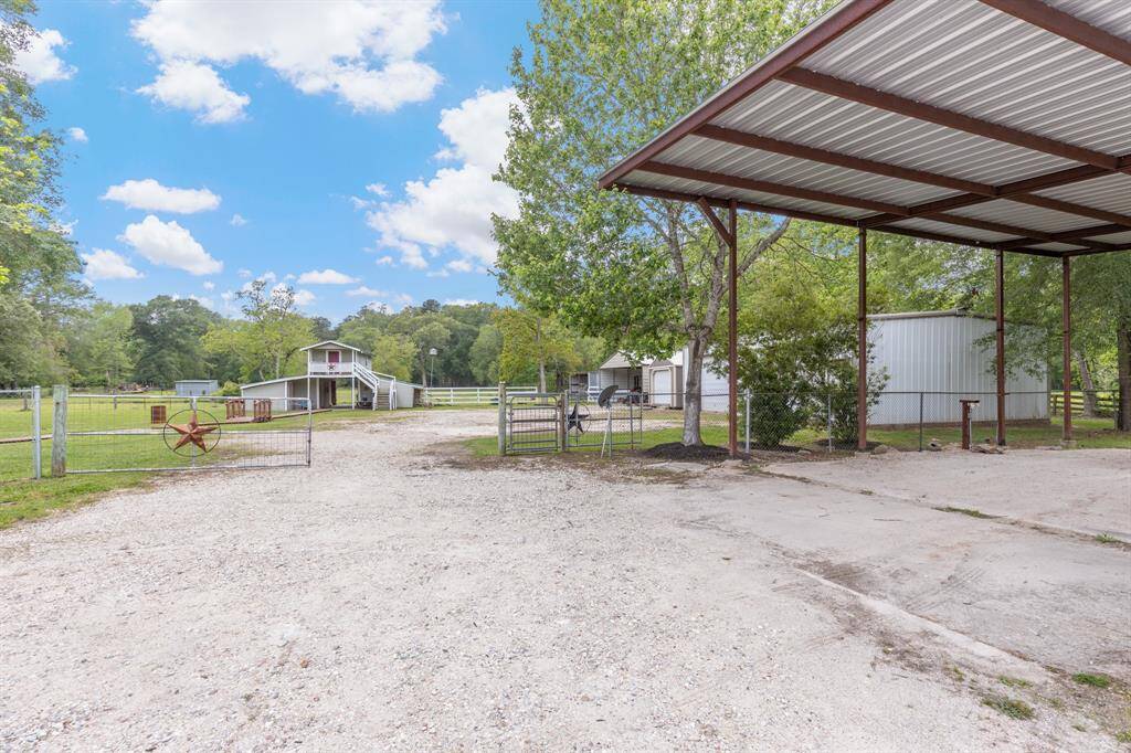 Spacious rural property with a large metal carport, gravel driveway, and a barn. The area features open green spaces, fenced enclosures, and mature trees, ideal for outdoor activities and animal care.