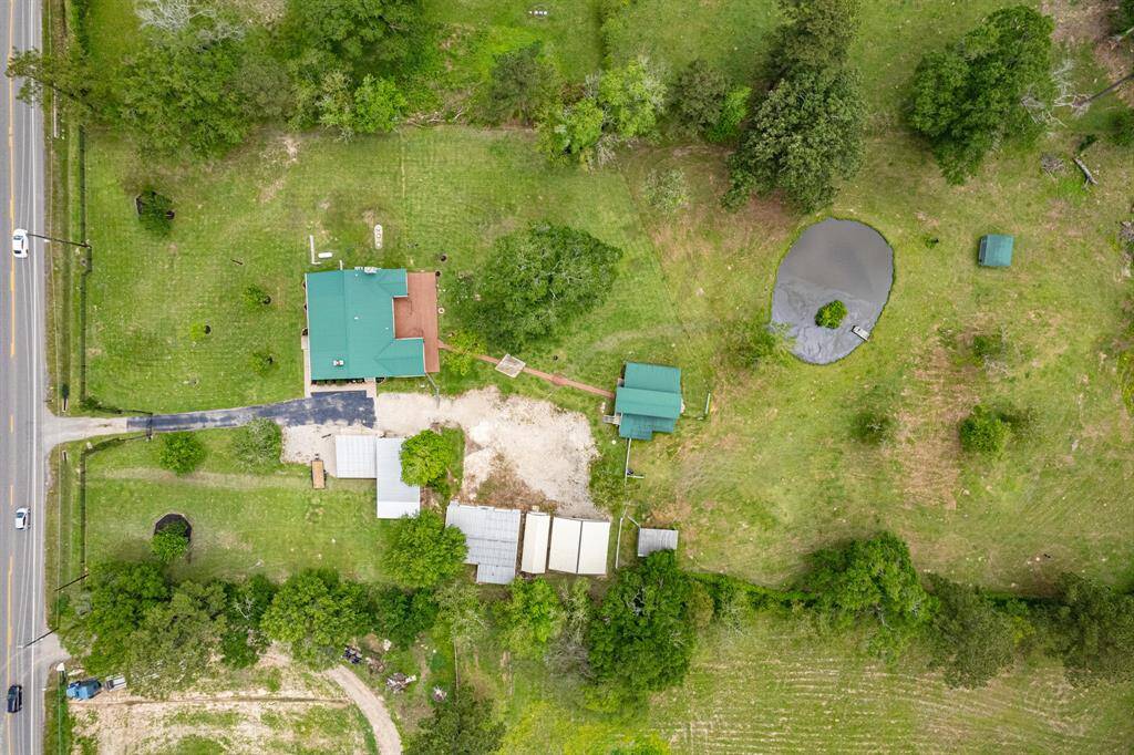 This aerial view shows a rural property with a green-roofed house, a driveway, and several outbuildings. The spacious yard includes a pond, trees, and open grassy areas, offering privacy and a scenic countryside setting.