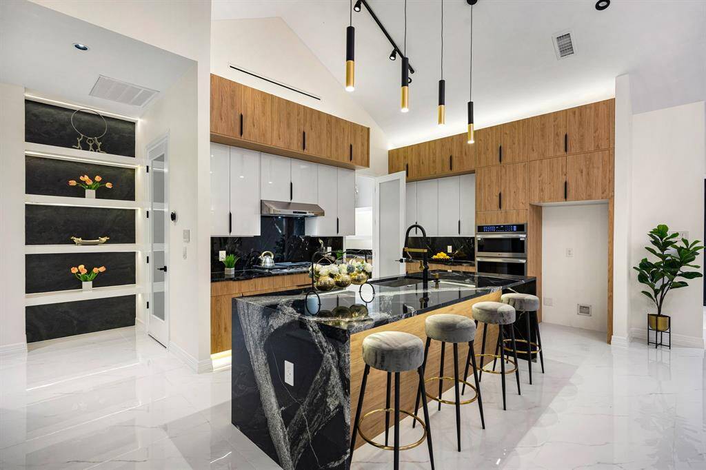 Step into this stunning modern kitchen featuring sleek high-gloss soft close cabinetry, warm wood finishes, and elegant black marble countertops. The spacious island offers ample workspace, perfectly complemented by dramatic pendant lighting and a vaulted ceiling that enhances the open, airy feel. High-end built-in appliances and under-cabinet lighting add both functionality and style, while the polished marble floors tie the space together with a luxurious touch.
