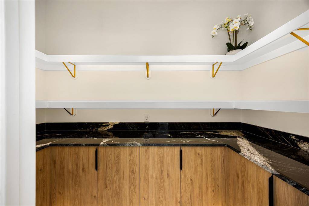 This spacious walk-in pantry offers both style and functionality with custom wood cabinetry, striking black stone countertops, and elegant veining that elevates the design. Open shelving with gold-accented brackets provides ample storage and display space, while under-shelf lighting potential enhances both visibility and ambiance. A perfect blend of luxury and practicality for any modern kitchen.