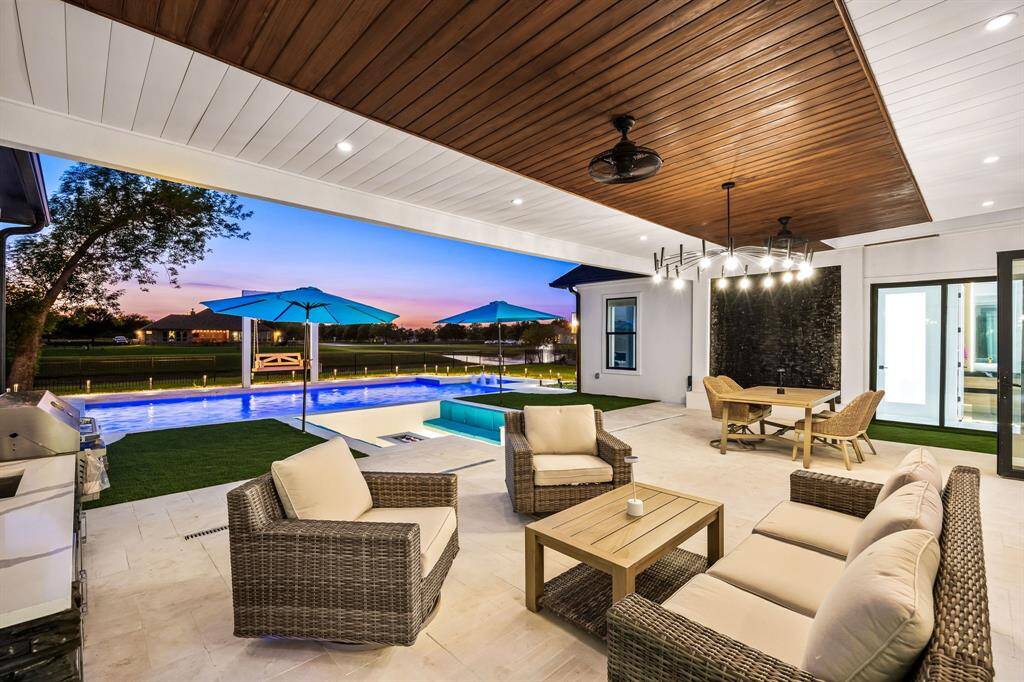 This stunning outdoor oasis boasts a luxurious covered patio with rich wood-paneled ceiling accents, recessed lighting, and ceiling fans for year-round comfort. A sleek built-in grill and prep area enhance the convenience of the outdoor kitchen. Overlooking a contemporary pool with vibrant lighting, spa, and in-pool lounge, the space is framed by open views and elegant landscaping—perfect for relaxing or entertaining in style.